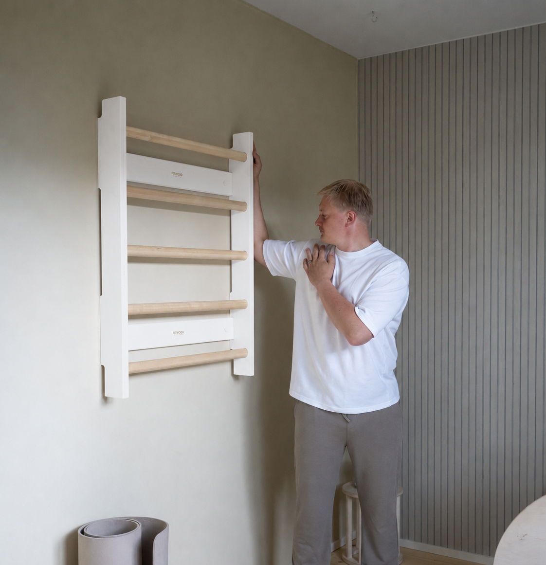 A man in a white t-shirt and gray sweatpants stands next to a wall-mounted wooden ladder shelf in a modern room with beige walls and wood flooring.
