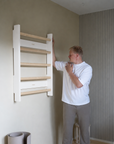 A man in a white t-shirt and gray sweatpants stands next to a wall-mounted wooden ladder shelf in a modern room with beige walls and wood flooring.